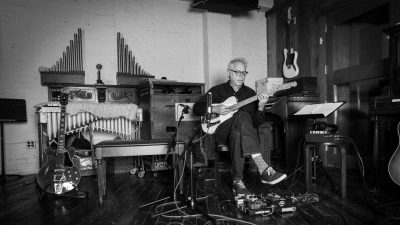 Jazz guitarist Bill Frisell, a white man with dark framed glasses and white hair, sits on a chair and plays his guitar while surrounded by various musical instruments and stands in this black and white photo.
