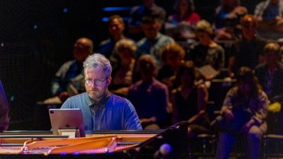 Pianist Conor Hanick, a middle aged white man, performs on stage in front of an audience while reading music from an iPad.