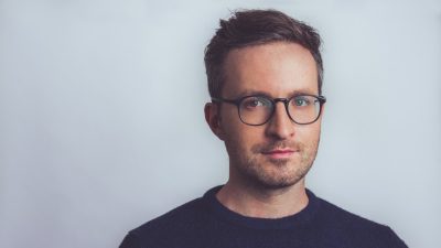 Pianist Conor Hanick, a middle aged white man, wears dark rimmed glasses and a dark shirt in this headshot.