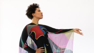 Portraits of vocalist Julia Bullock, 21C Media Group Inc. Bullock, a young Black woman, wears a multicolored patterned dress with dramatic gauzy sleeves.