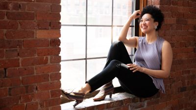 Portraits of vocalist Julia Bullock, 21C Media Group Inc. Bullock, a young Black woman, wears a light purple top and black pants and sits in the brick sill of a window