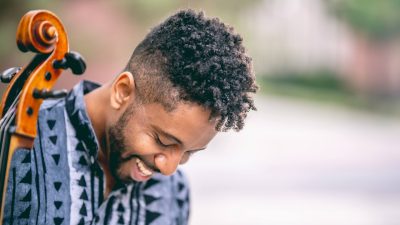 Cellist Seth Parker Woods, a young Black man, holds his cello outdoors in a city park, wearing a grey patterned button down tunic.