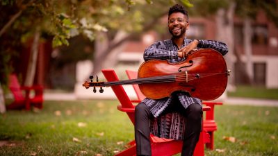 Cellist Seth Parker Woods, a young Black man, holds his cello outdoors in a city park while sitting on the arm of a bright red Adirondack chair, wearing a grey patterned button down tunic.