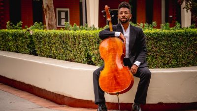Cellist Seth Parker Woods, a young Black man, holds his cello outdoors in a city park, wearing a suit.
