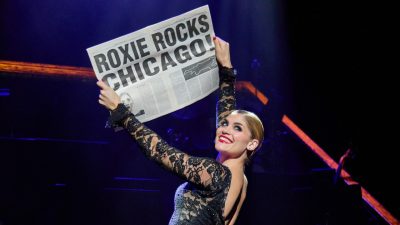 Roxie Hart stands with her back to the audience and holds up a newspaper with a headline that reads "Roxie Rocks Chicago" on stage during a performance of "Chicago."