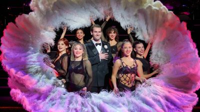 Cast of "Chicago," a white man in a tuxedo surrounded by cabaret performers in black costumes with large white and pink feather fans, perform on stage.