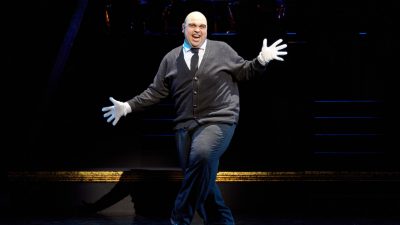A cast member of "Chicago" performs a solo number on stage. The man is white, balding, and heavyset and wears a cardigan and grey pants.
