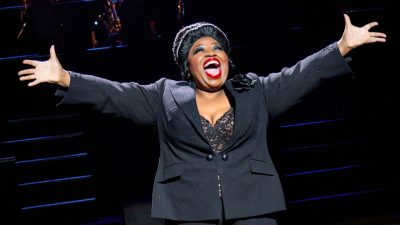 A cast member of "Chicago" performs a solo number on stage. The woman is Black and wears a black suit, lacy top, and red lipstick, her arms outspread while she hits a note.