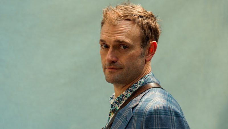 Mandolinist Chris Thile stands in front of a sea glass green background in a plaid jacket.