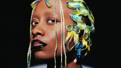 Jazz singer Cécile McLorin Salvant, a middle aged Black woman, wears a multicolored wig and black lipstick and stares down the camera in front of a black background.