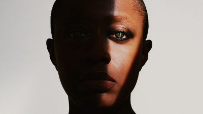 Jazz singer Cécile McLorin Salvant, a middle aged Black woman, wears snake eye contact lenses, only a slice of her face illuminated, in front of a white background.