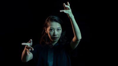 Conductor Miho Hazama, a young Asian woman, gestures towards the camera as if conducting, lit under a dim green light in front of a black background.
