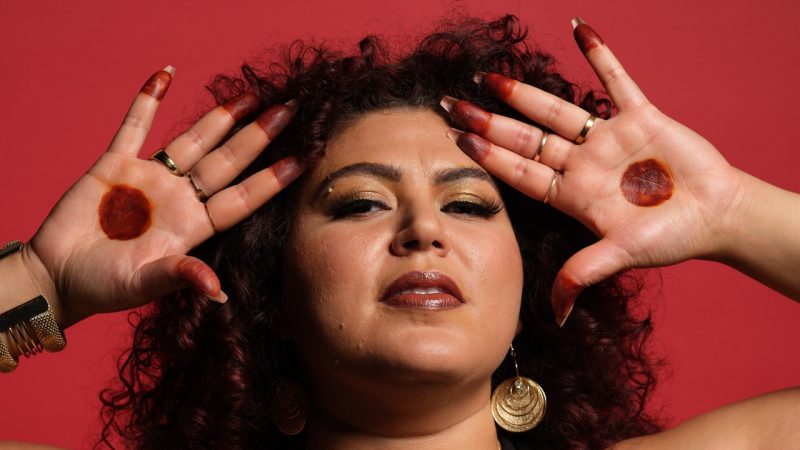 2025-2026 season Moss Arts Center guest curator Esraa Warda, an Algerian American dance performer with long curly dark brown hair. She wear gold earrings and rings and shows the palms of her hands to the camera, each fingertip and a circle in the center of each palm painted red.