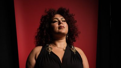 2025-2026 season Moss Arts Center guest curator Esraa Warda, an Algerian American dance performer with long curly dark brown hair. She wear gold earrings and a black jumpsuit and sits in front of a red backdrop.