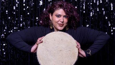 Guest curator Esraa Warda holds a hand drum in front of a sequined curtain.