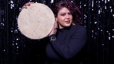 Guest curator Esraa Warda holds a hand drum in front of a sequined curtain.