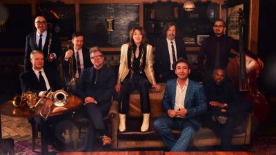 The members of New York jazz ensemble the Hot Sardines gathers in a jazz club, some holding their instruments.