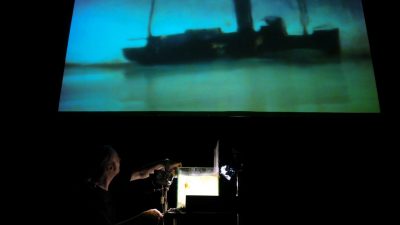 Members of Hotel Modern perform "The Great War" using an overhead projector and a large screen showing a U-boat.
