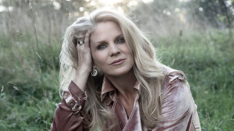 Broadway and screen actress Kelli O'Hara, a middle aged blonde woman, sits in a grassy field in the early morning wearing a dusty rose velvet dress.