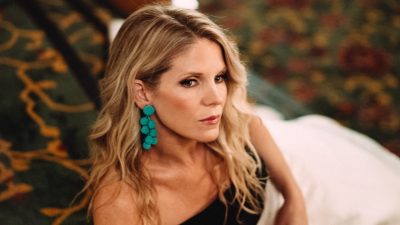 Broadway and screen actress Kelli O'Hara, a middle aged blonde woman, wears a black, red, and white dress and turquoise earrings and reclines on the carpeted steps in a performing arts center.