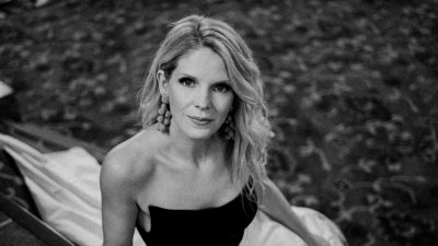 Broadway and screen actress Kelli O'Hara, a middle aged blonde woman, wears a black and white dress and reclines on the carpeted steps in a performing arts center in this black and white image.