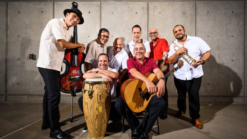 The members of Kiki Valera y su Son Cubano sit and stand together holding their instruments.