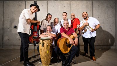 The members of Kiki Valera y su Son Cubano sit and stand together holding their instruments.