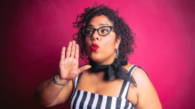 La Marisoul, the lead singer of La Santa Cecilia, a Latina woman in a black and white striped dress in front of a red background.