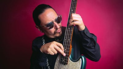 A member of La Santa Cecilia plays his bass guitar in front of a red background.