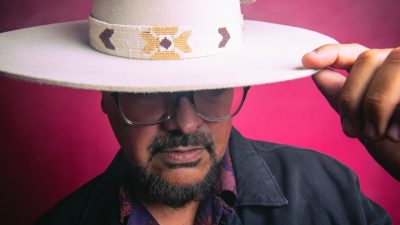 A member of La Santa Cecilia tips his wide-brimmed white hat in front of a red background.