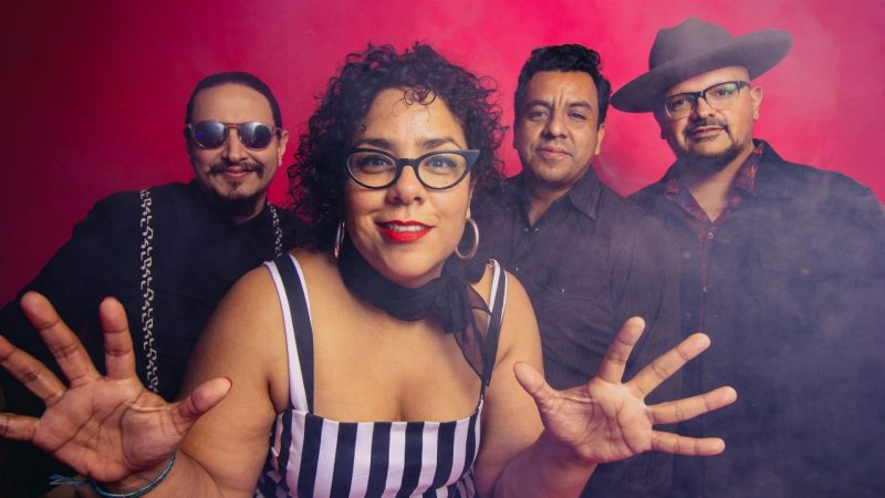 The members of La Santa Cecilia stand in front of a red background, some theatrical smoke in the frame. The lead singer, Marisoul, stands towards the front in black framed cat eye glasses, a black and white striped dress, and a black scarf, her hands spread wide.