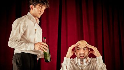 Physical theatre due Bert and Nasi perform "L'Addition" in front of a red velvet curtain. One man sits with his head in his hands behind a wine glass, pulling a face, while the other approaches with a bottle of wine.