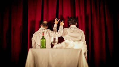 Physical theatre due Bert and Nasi perform "L'Addition" in front of a red velvet curtain. Both sit at a table with their backs to the camera and lift their arms up as if cheersing with imaginary glasses.