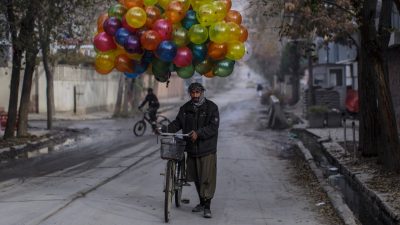 A middle eastern man walks a bike wearing a dark jacket and green pants. He holds a cluster of brightly colored balloons overhead. Photo by National Geographic Explorer Muhammed Muheisen.