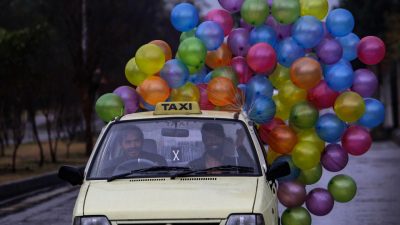 Two men ride in a small yellow taxi, the passenger smiling widely and holding on the strings of a larger cluster of multicolored balloons, which trail behind the taxi. Photo by National Geographic Explorer Muhammed Muheisen.