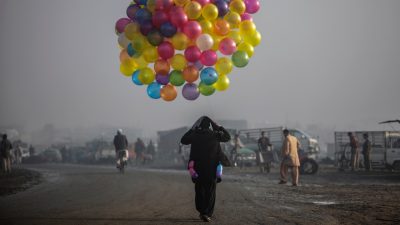 A man in a middle eastern town, wearing all black, walks through the street on a hazy morning, holding onto a large cluster of multicolored balloons. Photo by National Geographic Explorer Muhammed Muheisen.