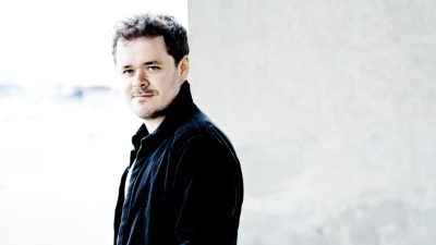 Pianist Benjamin Grosvener, a white man with brown hair and blue eyes, stands near a marble exterior wall.
