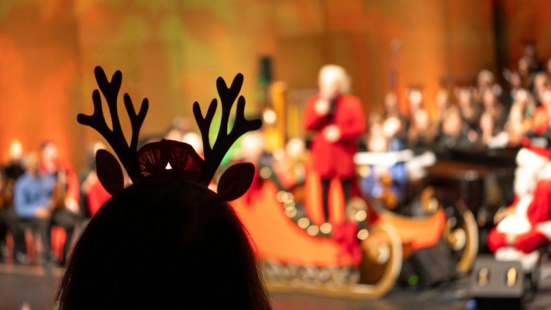 A woman wearing reindeer antlers watches the Roanoke Symphony Orchestra's "Holiday Pops Spectacular" as conductor David Stewart Wiley speaks and Santa looks on.