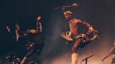 A member of Step Afrika! wears a traditional African garment and performs animatedly on stage.