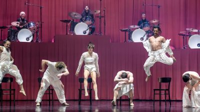 Members of Stephanie Lake Company wear white costumes and dance, some leaping into the air, on stage in front of a towering pink curtain and several drummers.