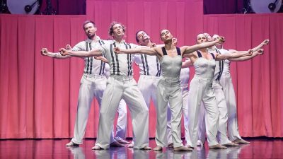 Members of Stephanie Lake Company wear white costumes and perform on stage in front of a towering pink curtain and several drummers.