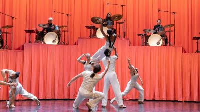 Members of Stephanie Lake Company wear white costumes and perform on stage in front of a towering pink curtain and several drummers.