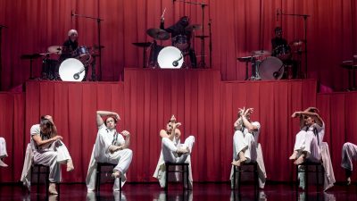 Members of Stephanie Lake Company wear white costumes and perform on stage in front of a towering pink curtain and several drummers.
