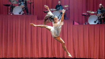 Members of Stephanie Lake Company wear white costumes and perform on stage in front of a towering pink curtain and several drummers.