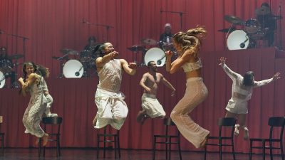 Members of Stephanie Lake Company wear white costumes and perform on stage in front of a towering pink curtain and several drummers.