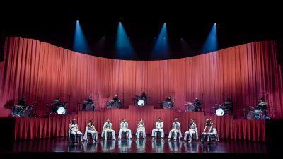 Members of Stephanie Lake Company wear white costumes and perform on stage in front of a towering pink curtain and several drummers.