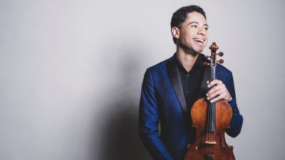 Jamaican-American violist Jordan Bak, a young man with short dark brown hair, wears a navy suit and holds his viola.