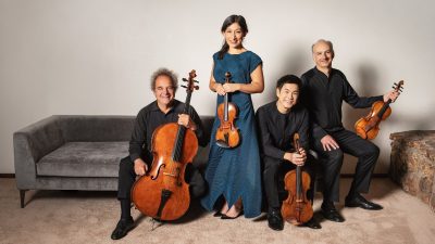 The Takács Quartet in concert attire hold their instruments in a living room with a grey couch, carpet, and a stone hearth.