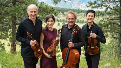 The Takács Quartet in concert attire in a grassy field holding their instruments. Trees and a mountain range are visible behind them.