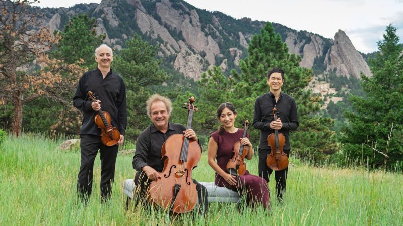The Takács Quartet in concert attire in a grassy field holding their instruments. Trees and a mountain range are visible behind them.